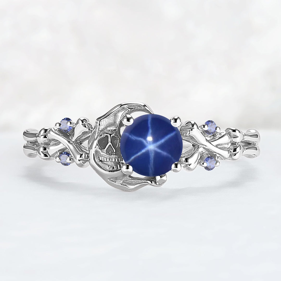 A  ring featuring a central round blue star ruby gemstone, flanked by a detailed skull motif and small round blue accent stones, set against a soft white background.