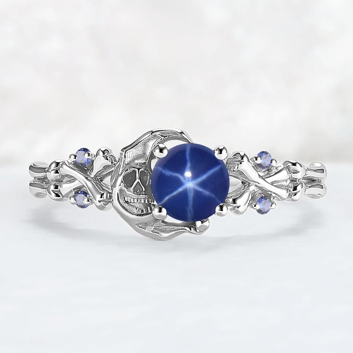 A  ring featuring a central round blue star ruby gemstone, flanked by a detailed skull motif and small round blue accent stones, set against a soft white background.